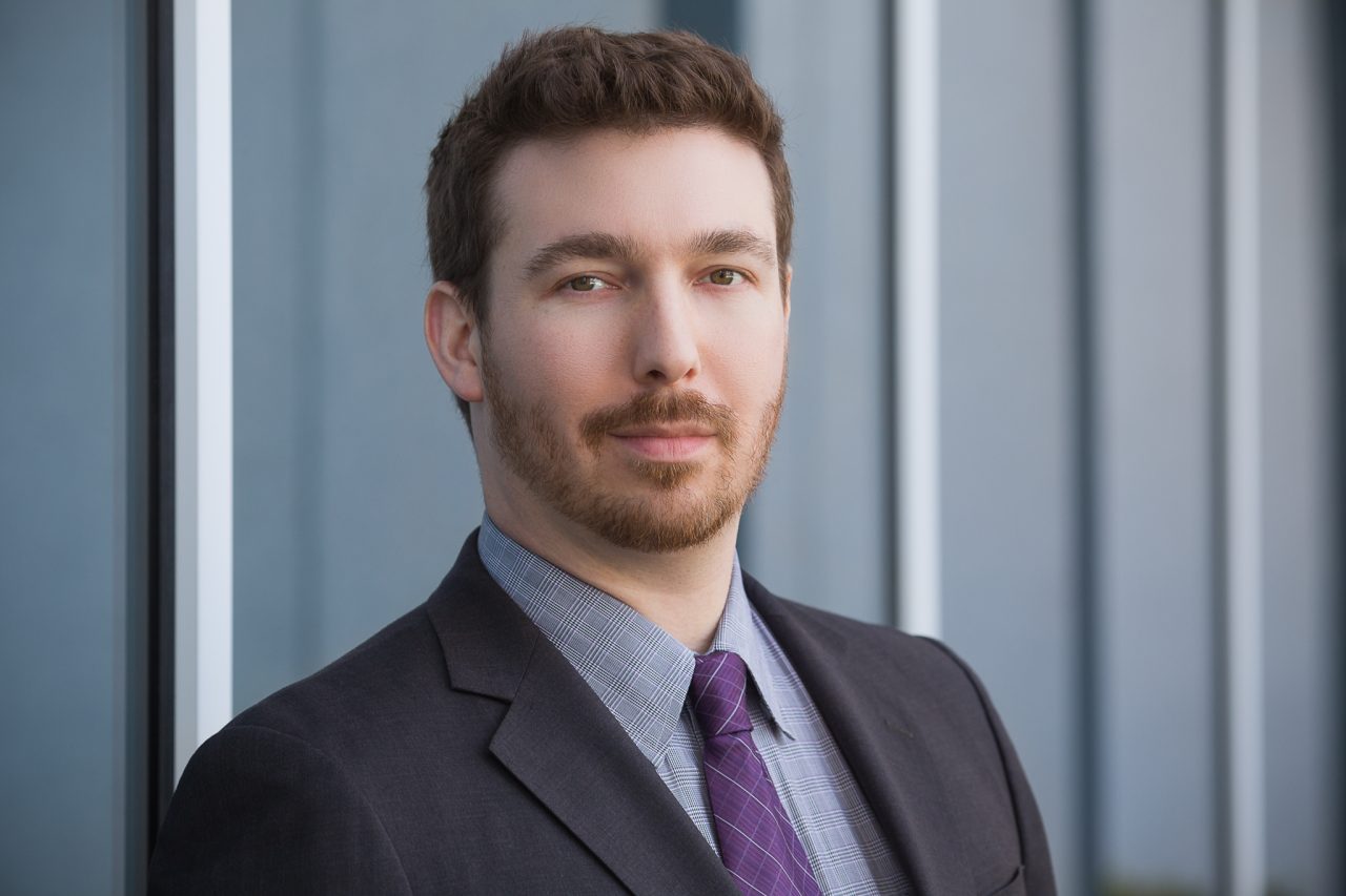 Professional business headshot of a man in a suit and tie, posing outdoors against a modern building.