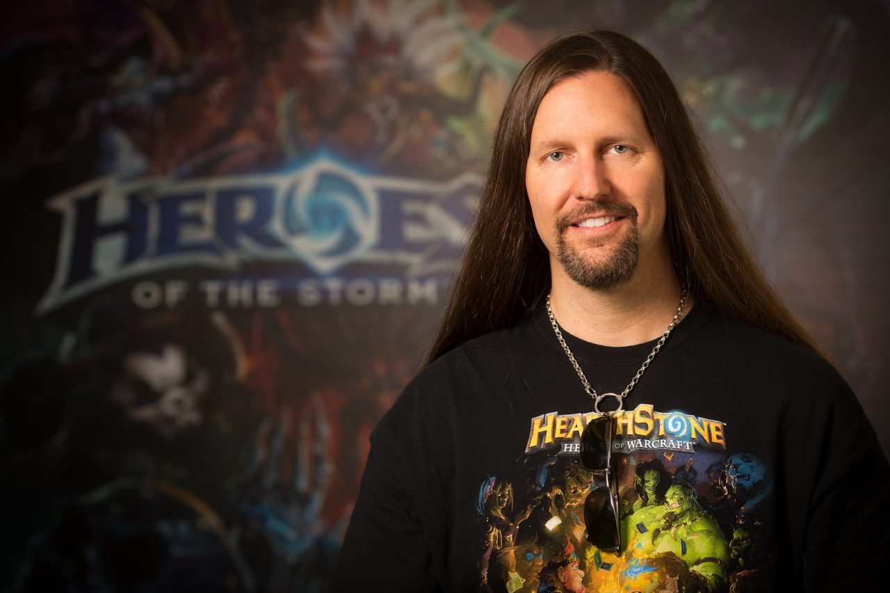 Professional headshot of a person standing in front of a blurred Heroes of the Storm promotional backdrop.