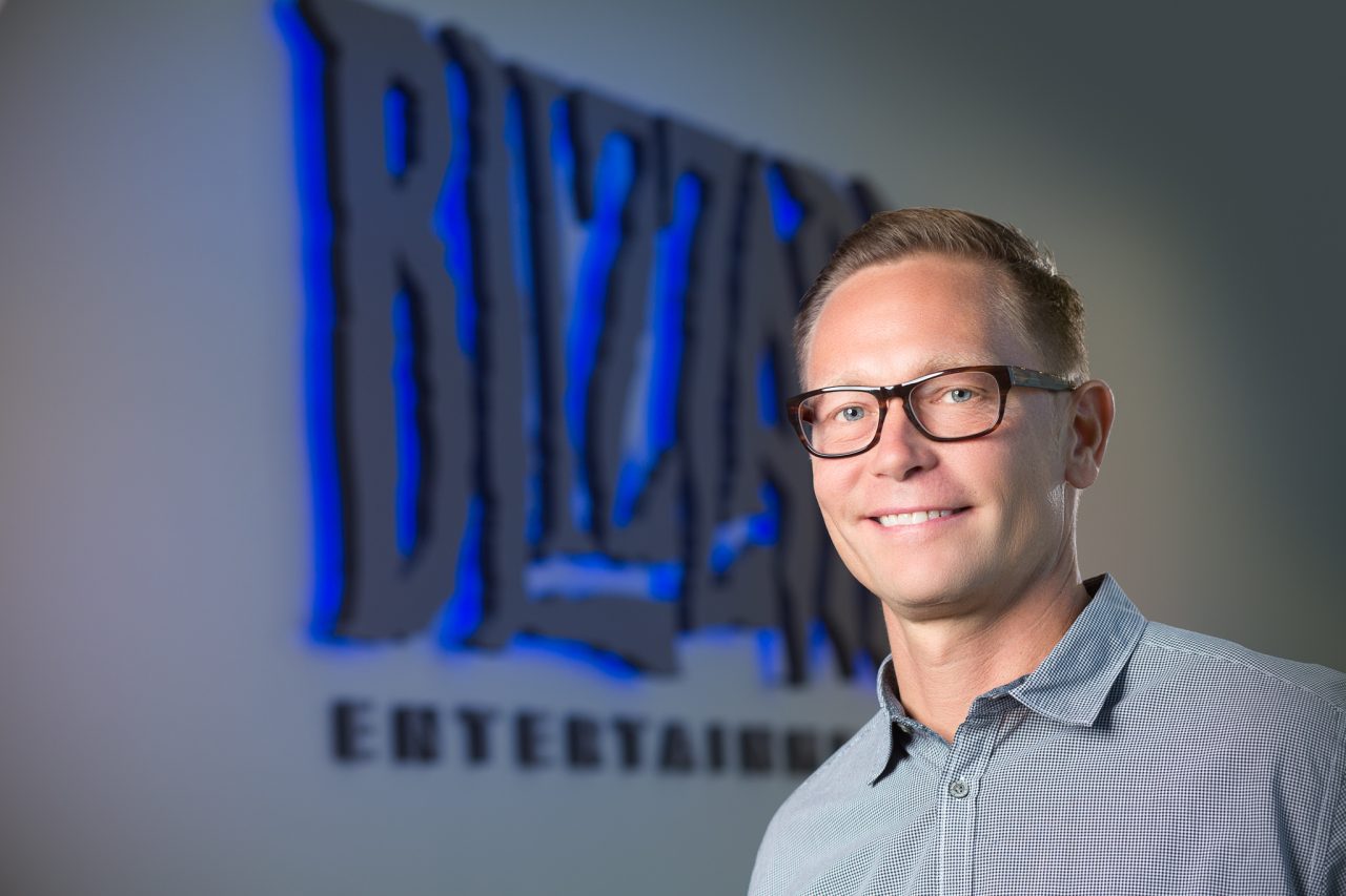 Professional headshot of a person in glasses standing in front of a blurred Blizzard Entertainment logo with blue lighting.