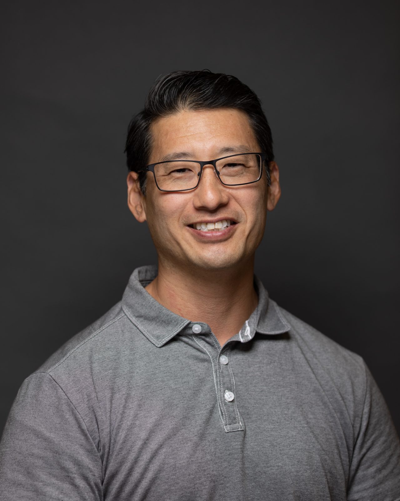 A person wearing glasses and a gray collared shirt smiles in a studio-style corporate portrait.