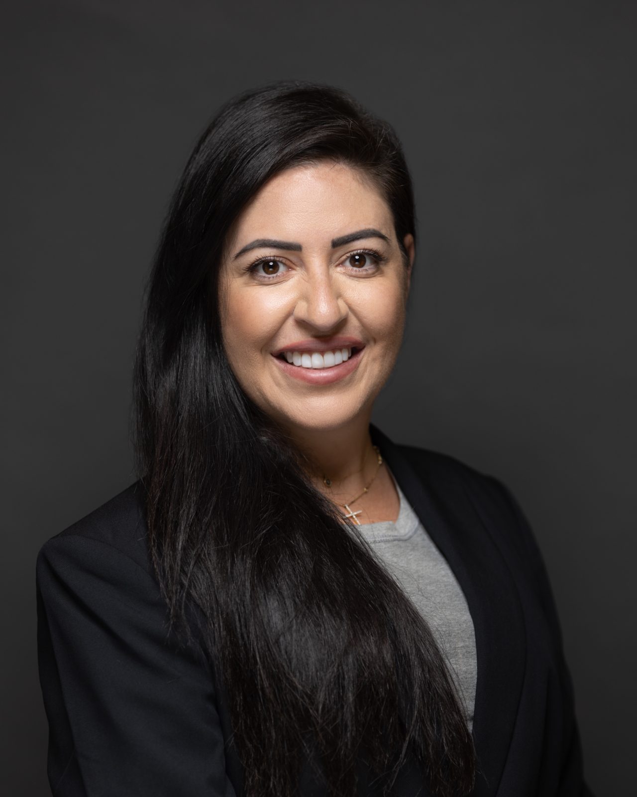 A person with long dark hair wearing a blazer smiles in a professional studio headshot.