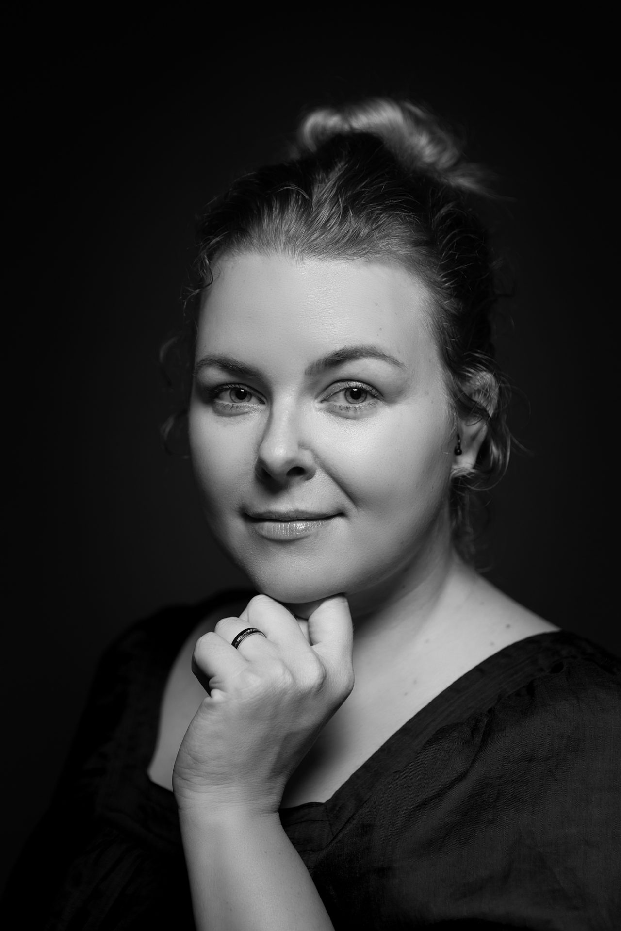 A person rests their hand near their chin and smiles softly in a black-and-white studio portrait.