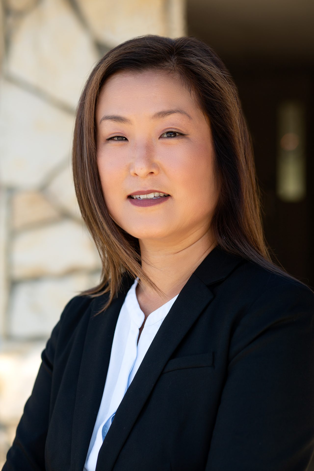 A real estate agent poses outdoors in professional attire, with a stone wall softly blurred in the background.